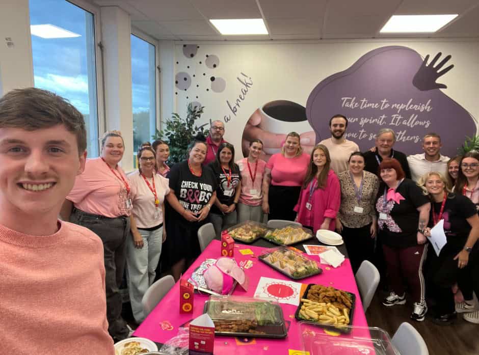 Breast Cancer Now fundraiser at HCL'S head office, with a group of individuals behind a buffet table all wearing pink, smiling and posing for the camera.