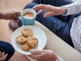 An image of a woman handing over a cup of tea and biscuits to an elderly man sat on a couch.