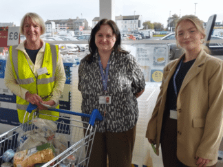 Picture of 3 women standing in the opening entrance of a supermarket, in front of a trolley full of donated goods.