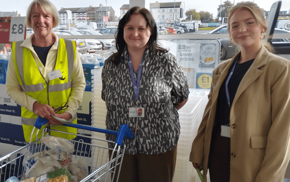 Picture of 3 women standing in the opening entrance of a supermarket, in front of a trolley full of donated goods.