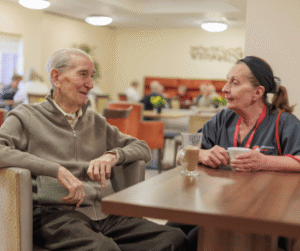 Image of two people sat at a table in a canteen setting, chatting over a cup of tea.