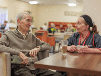 Image of two people sat at a table in a canteen setting, chatting over a cup of tea.