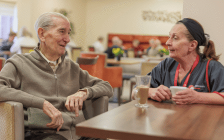 Image of two people sat at a table in a canteen setting, chatting over a cup of tea.