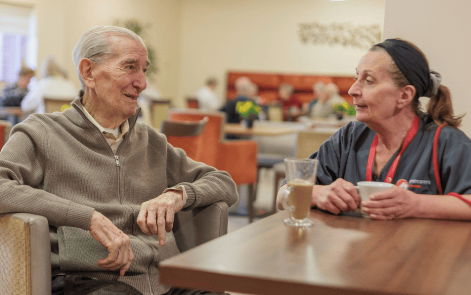 Image of two people sat at a table in a canteen setting, chatting over a cup of tea.