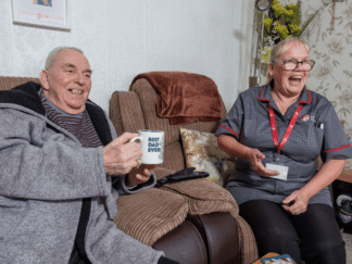 2 people in a home setting, sitting on a couch with a cup of tea laughing.