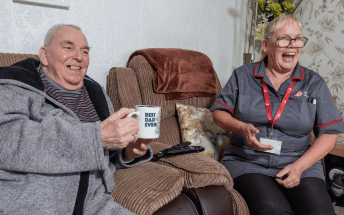 2 people in a home setting, sitting on a couch with a cup of tea laughing.