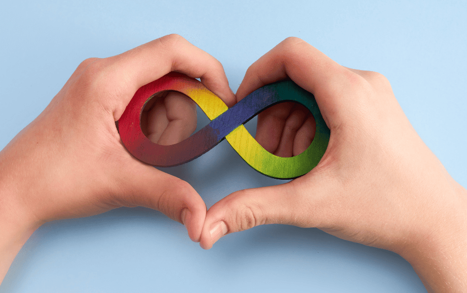 Image of a pair of hands in the shape of a love heart, holding a rainbow coloured infinity sign against a blue background.