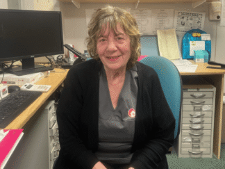 Image of a woman sat in an office setting on a chair, smiling at the camera wearing a care worker uniform.