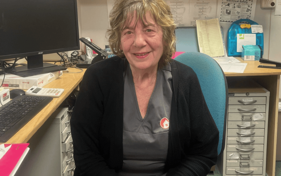 Image of a woman sat in an office setting on a chair, smiling at the camera wearing a care worker uniform.