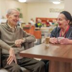 Image of two people sat at a table in a canteen setting, chatting over a cup of tea.