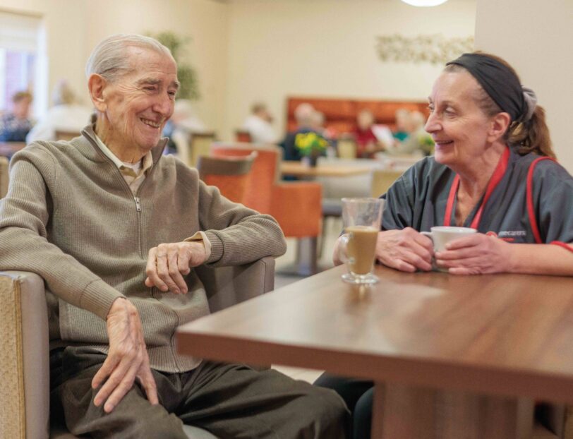 Image of two people sat at a table in a canteen setting, chatting over a cup of tea.