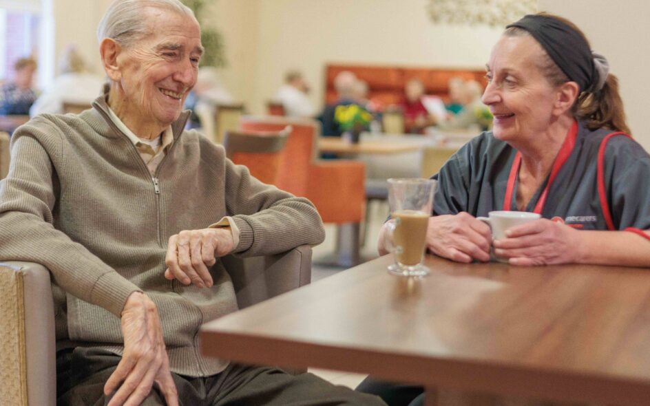 Image of two people sat at a table in a canteen setting, chatting over a cup of tea, highlighting companionship services.