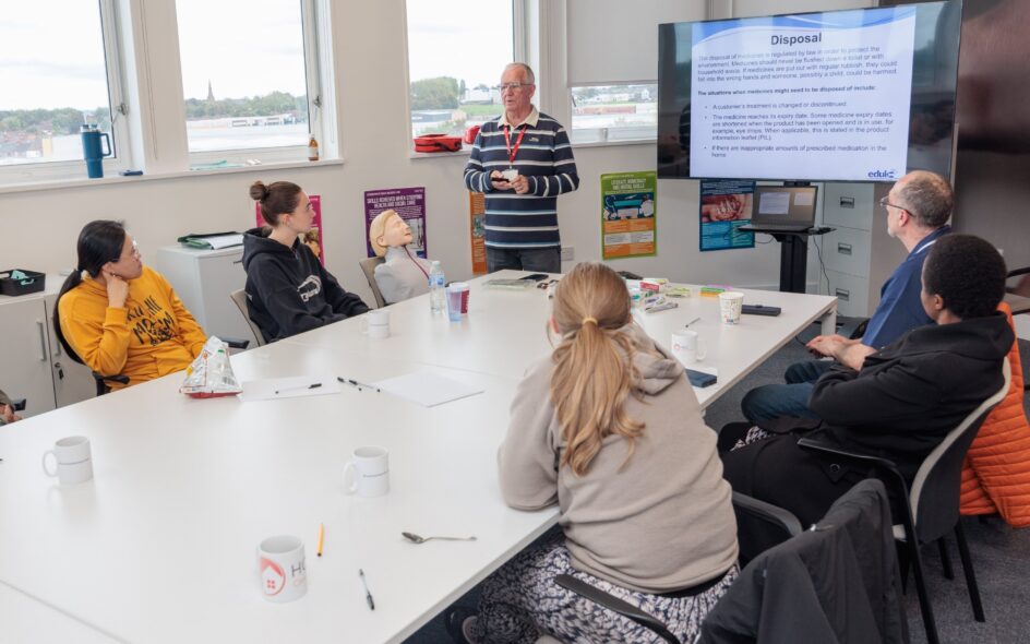 Group of people in a classroom, with a trainer at the head of the table, leading a PowerPoint presentation. first year in care