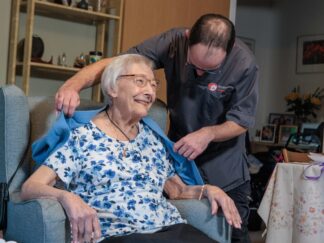 An image of a Care Assistant and an elderly lady, helping put a jacket over her.