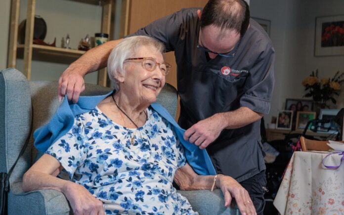 An image of a Care Assistant and an elderly lady, helping put a jacket over her.