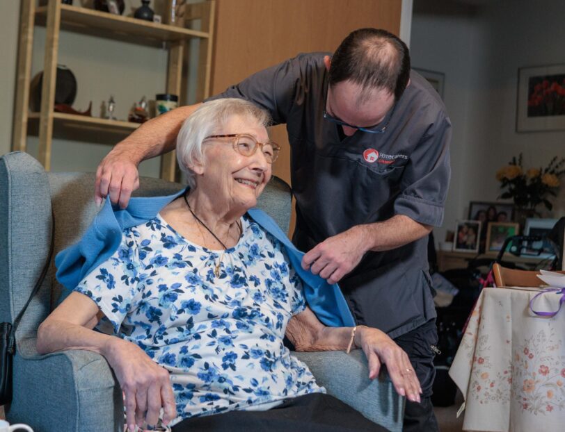 An image of a Care Assistant and an elderly lady, helping put a jacket over her.