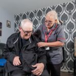 An image of a Care Assistant and an elderly man, helping put a jacket over him