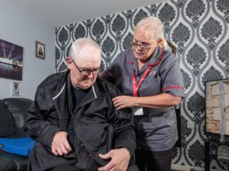 An image of a Care Assistant and an elderly man, helping put a jacket over him