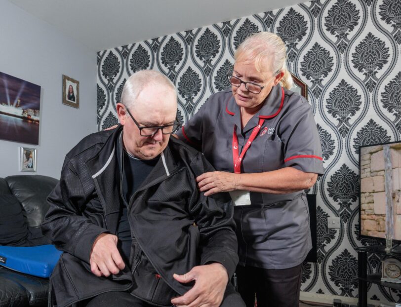 An image of a Care Assistant and an elderly man, helping put a jacket over him