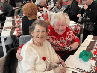 An image of 2 elderly women sat at a table, enjoying a Christmas party.