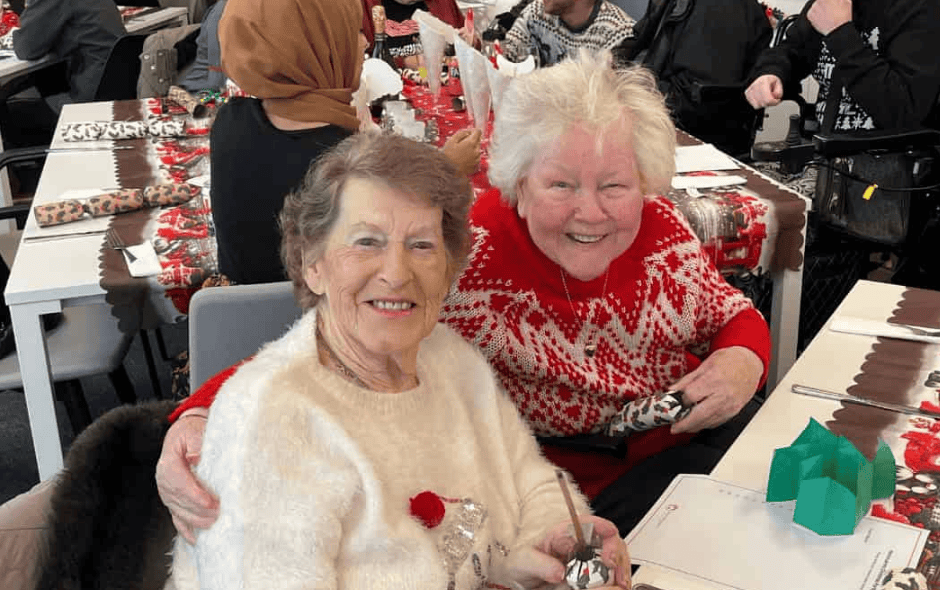 An image of 2 elderly women sat at a table, enjoying a Christmas party.
