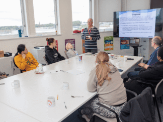 Group of people in a classroom, with a trainer at the head of the table, leading a powerpoint.