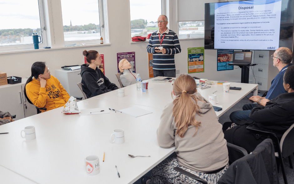 Group of people in a classroom, with a trainer at the head of the table, leading a powerpoint.