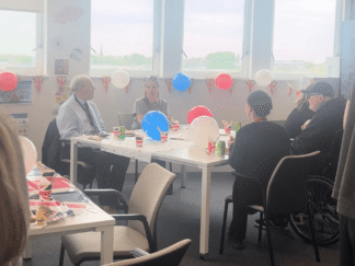 An image of people sat at an afternoon tea party chatting, with red, white and blue decorations in the background.