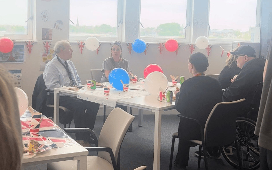 An image of people sat at an afternoon tea party chatting, with red, white and blue decorations in the background.