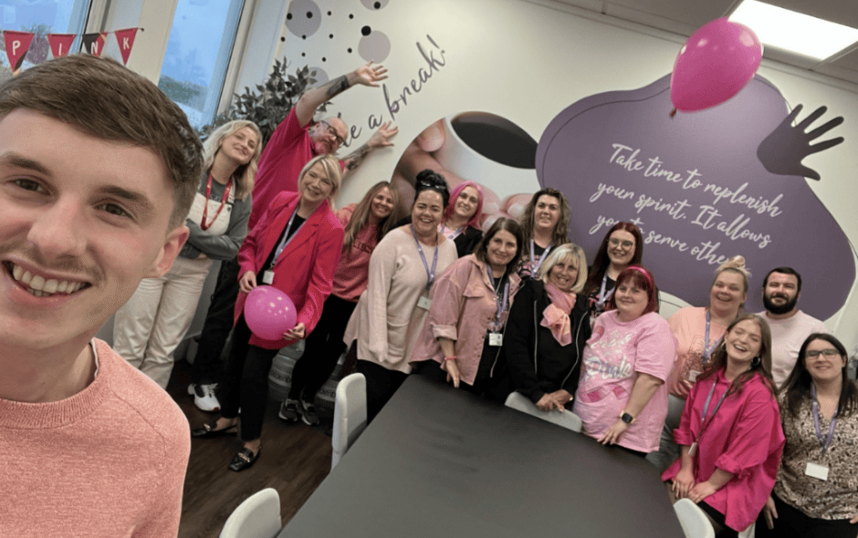 Group of colleagues in a kitchen area all wearing pink for a fundraiser, smiling for a selfie.