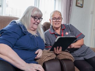 An image of an elderly woman and a care assistant in a grey and red uniform sat on a couch in a home setting, looking at a tablet screen. choosing care at home