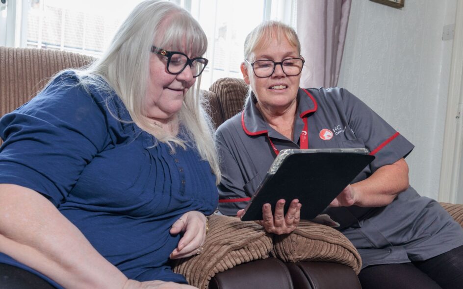 An image of an elderly woman and a care assistant in a grey and red uniform sat on a couch in a home setting, looking at a tablet screen. choosing care at home