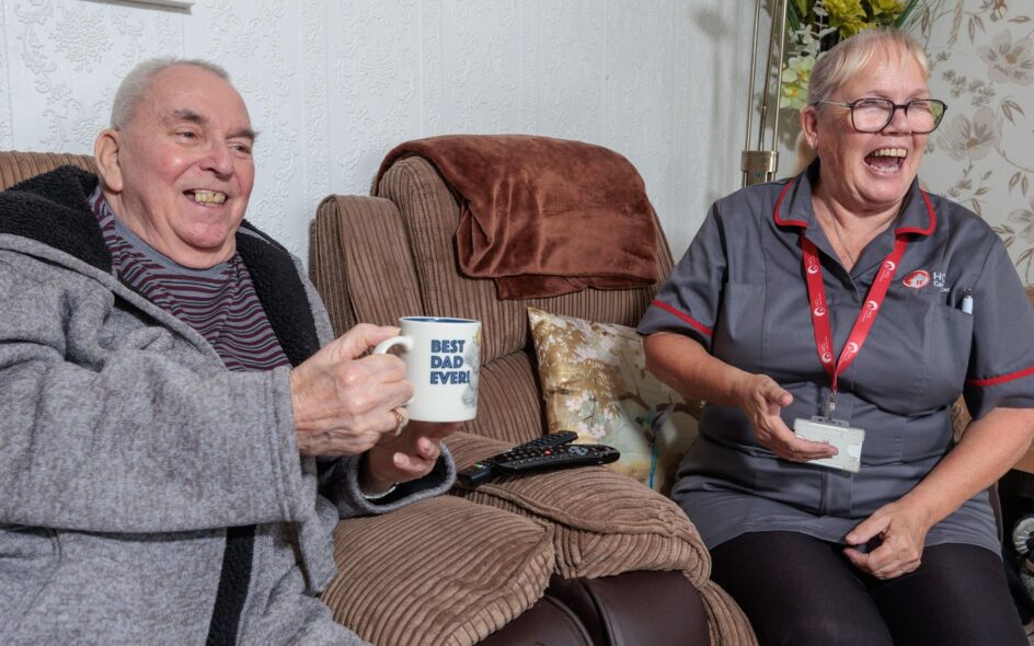 Image of a service user and a Care Assistant in a grey and red uniform, smiling and laughing whilst holding a cup of tea. services