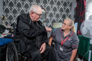 An image of a man in a wheelchair pointing at a woman in a grey and red care assistant uniform.