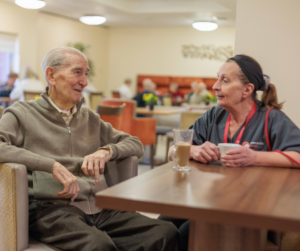 An image of an elderly man and a woman in a grey and red care assistant uniform sat in a cafe, sharing a cup of coffee.