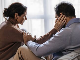 An image of a woman and a man where the woman is comforting the man by placing her hand on his face. Both are in a lightly lit room.