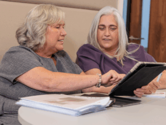 A man and a woman in an office setting taking part in a meeting, smiling whilst looking at a tablet.